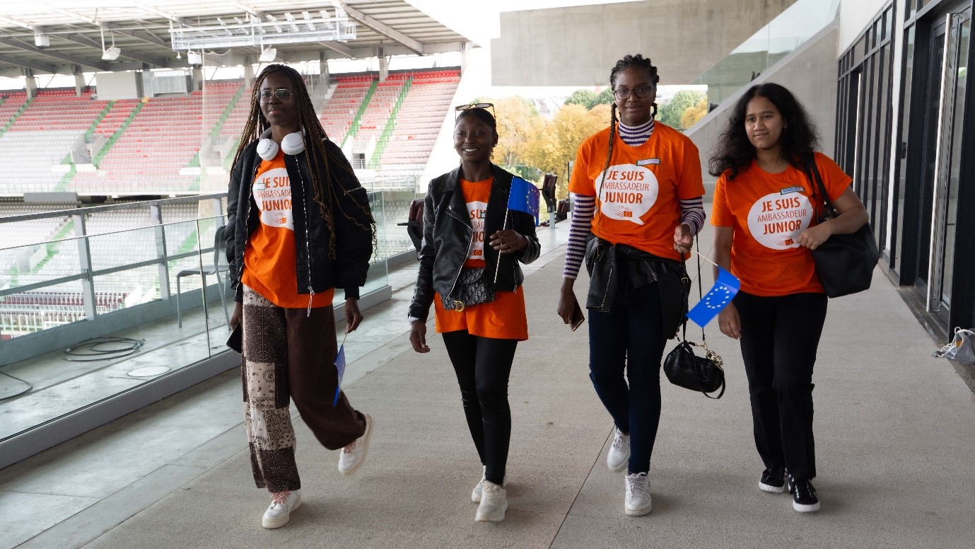 4 lycéennes portant des t-shirts orange sur lesquels il est écrit « je suis ambassadeur junior » et des drapeaux européens, marchent ensemble dans le stade, en avançant vers le photographe.